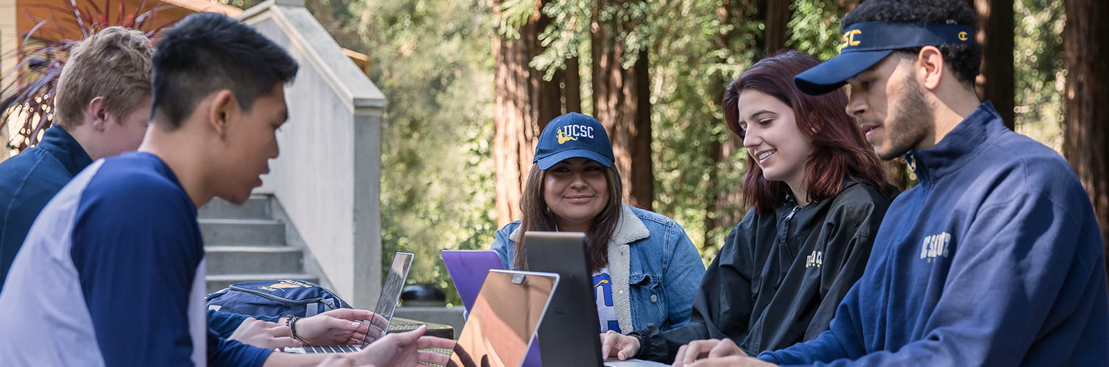 A group of five students sit using their laptops at a table in the redwoods. They are wearing UC Santa Cruz hats and jackets.