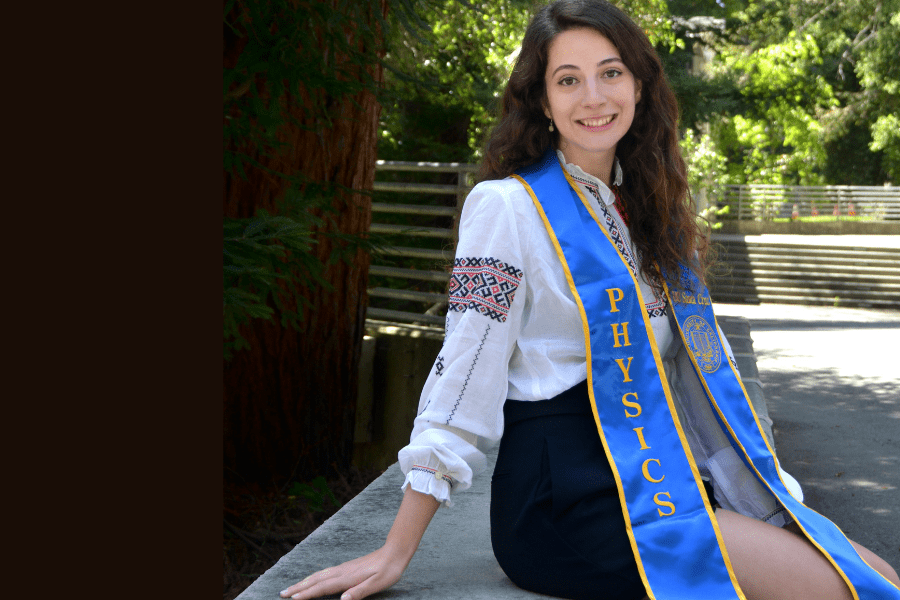 Portrait of Elizabeth Yunerman sitting on a concrete bench and wearing a graduation sash that reads "physics."