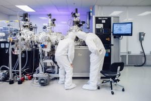 Two researchers in white full-body hazmat suits gaze into a piece of machinery in a lab.